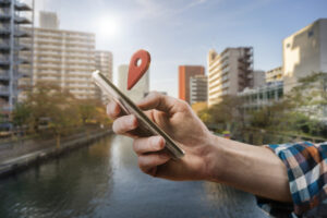 Une main tient un smartphone duquel sort une icône de géolocalisation, avec un paysage urbain et un canal en arrière-plan.