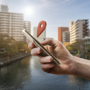 Une main tient un smartphone duquel sort une icône de géolocalisation, avec un paysage urbain et un canal en arrière-plan.